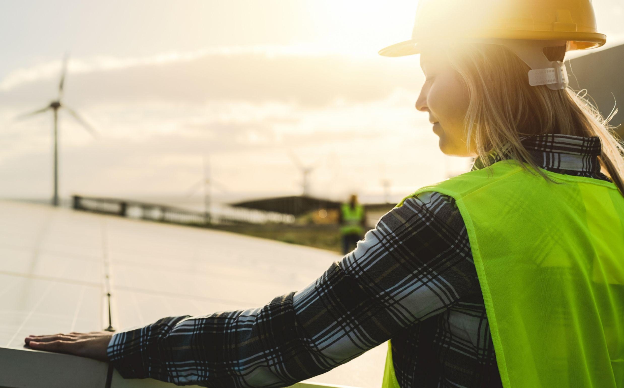 A person wearing a yellow hard hat and high-visibility green safety vest inspects solar panels at a solar farm during sunset. Wind turbines are visible in the background. The worker is wearing a plaid patterned long-sleeve shirt under their safety vest.