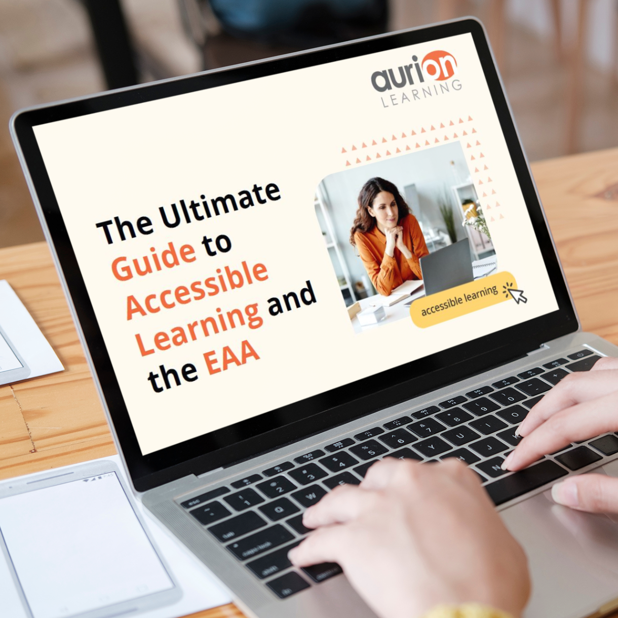 A laptop on a wooden table displaying a webpage from Aurion Learning. The screen shows a banner titled "The Ultimate Guide to Accessible Learning and the EAA" with "Guide," "Accessible Learning," and "EAA" highlighted in orange. The webpage features an image of a person with long dark hair wearing an orange top, seemingly in an office setting. There's also a yellow button labelled "accessible learning" with a cursor icon next to it.

A pair of hands is visible typing on the laptop keyboard. Next to the laptop is some papers or documents. The setting looks like a café or casual workspace with a warm wooden table surface. The Aurion Learning logo is visible at the top of the screen.