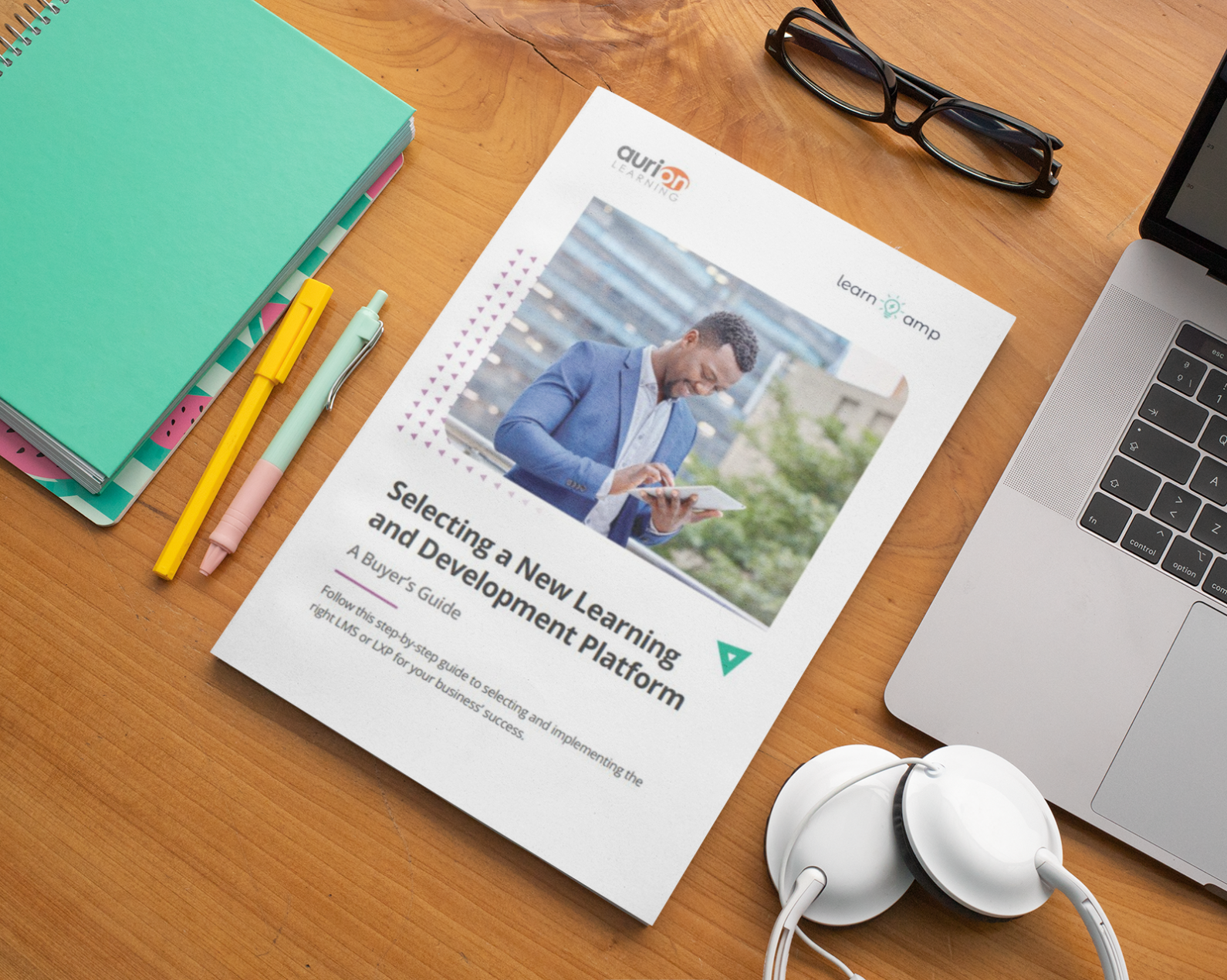 A professional workspace arranged on a wooden desk featuring a printed guide titled "Selecting a New Learning and Development Platform: A Buyer's Guide." The document is co-branded with Aurion Learning and Learn Amp logos, and includes an image of a man in business casual attire looking at a tablet outdoors.

The guide appears to be a comprehensive resource offering step-by-step guidance on selecting and implementing learning platforms for business success. 

Surrounding the document are typical workspace items including a turquoise spiral-bound notebook with colourful dividers, writing implements (a yellow pen and light blue pen), black-framed glasses, white earbuds, and a laptop visible at the edge of the frame.

The wooden desk surface and warm lighting create a professional yet approachable atmosphere, suggesting this is both a practical reference document and educational marketing material. The composition effectively presents the guide as a valuable resource for HR professionals, learning and development managers, or business decision-makers evaluating new training platforms for their organisations.