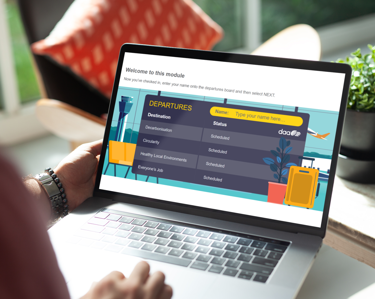 A laptop computer showing an interactive eLearning module with an airport departures board theme. The screen shows "Welcome to this module" with instructions to enter a name and select "NEXT." The central feature is a stylised departures board displaying various training topics including "Decarbonisation," "Circularity," "Healthy Local Environments," and "Everyone's Job," all showing "Scheduled" status. The interface includes a yellow name field showing "John" and features colourful airport-themed graphics including a control tower, luggage, and aircraft illustrations. The daa logo is visible in the top right corner. The laptop is positioned on a desk with a person's hands visible on the keyboard, wearing a dark beaded bracelet, suggesting an interactive learning environment focused on sustainability and workplace training.