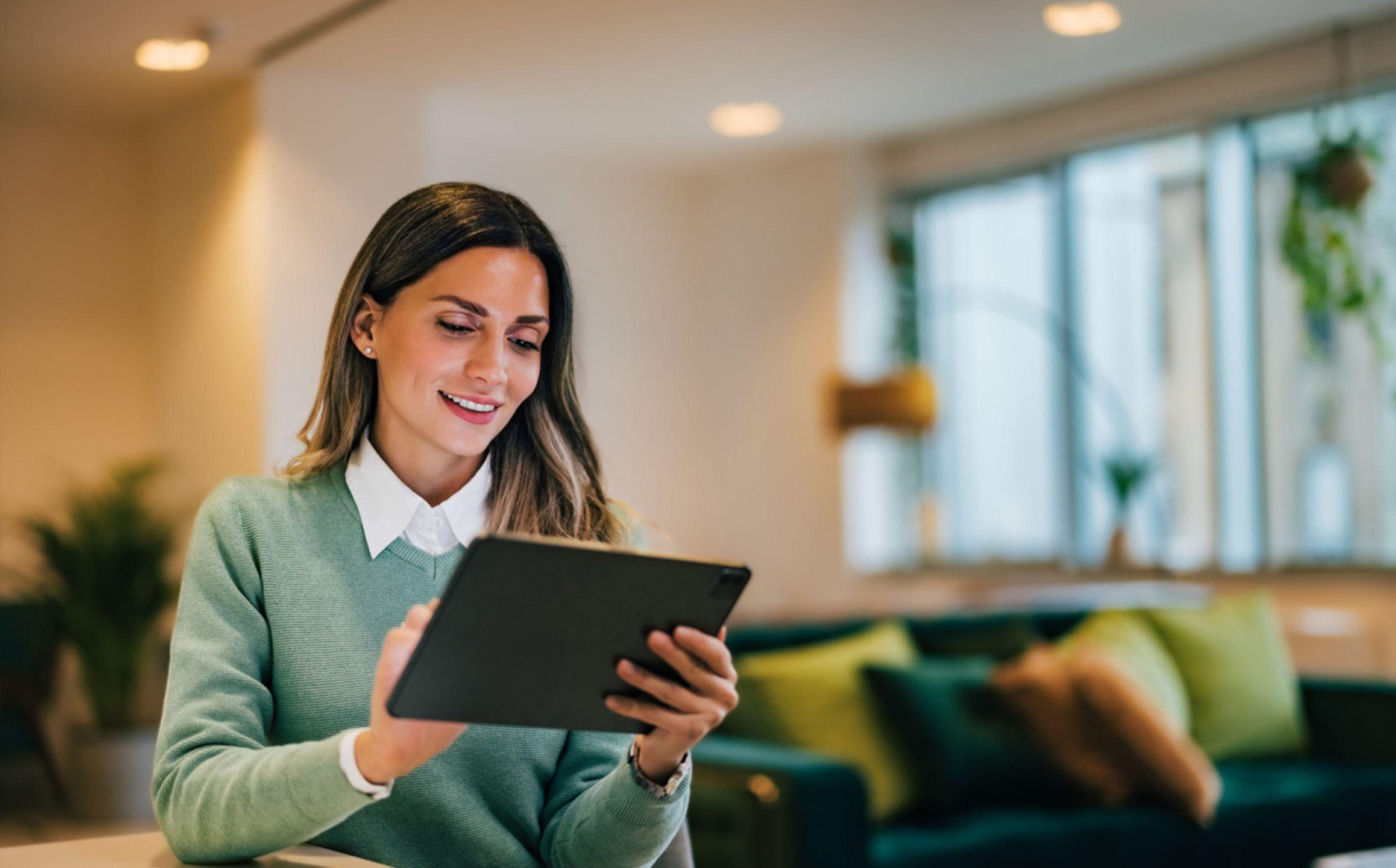 The image shows a woman in a green sweater sitting in a cozy-looking living room or workspace, using a tablet device with a focused expression. The background suggests a comfortable, welcoming environment with plants and other decor elements.