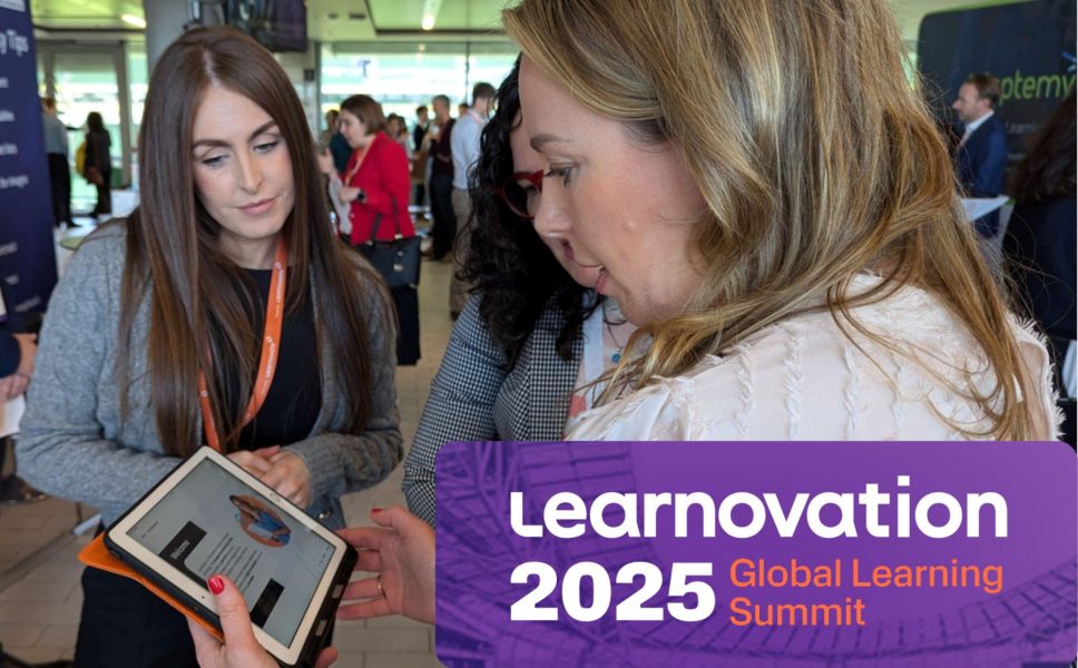 A photograph taken by Aurion Learning at Learnovation 2024. In the foreground, there are three women engaged in discussion - one with long brown hair wearing a grey jacket and orange lanyard looking at a tablet, another with blonde hair in a light-coloured top, and a third woman with dark hair wearing a checkered pattern outfit. The setting is a busy conference hall within Aviva Stadium, Dublin, with other attendees visible in the background.
The image includes branding for "Learnovation 2025 Global Learning Summit" with a purple banner overlay. The scene captures the collaborative, networking atmosphere typical of such professional events, with attendees sharing information and discussing content on digital devices.
