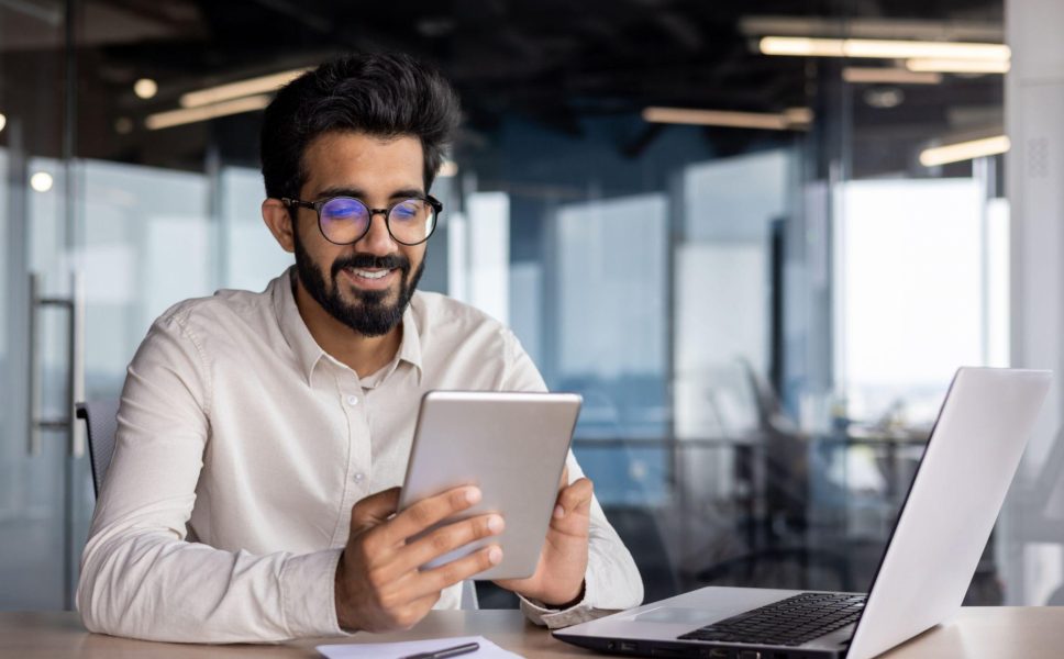 Smiling man wearing glasses and a white shirt sitting in a modern office, using a tablet with a laptop and notepad on the desk.