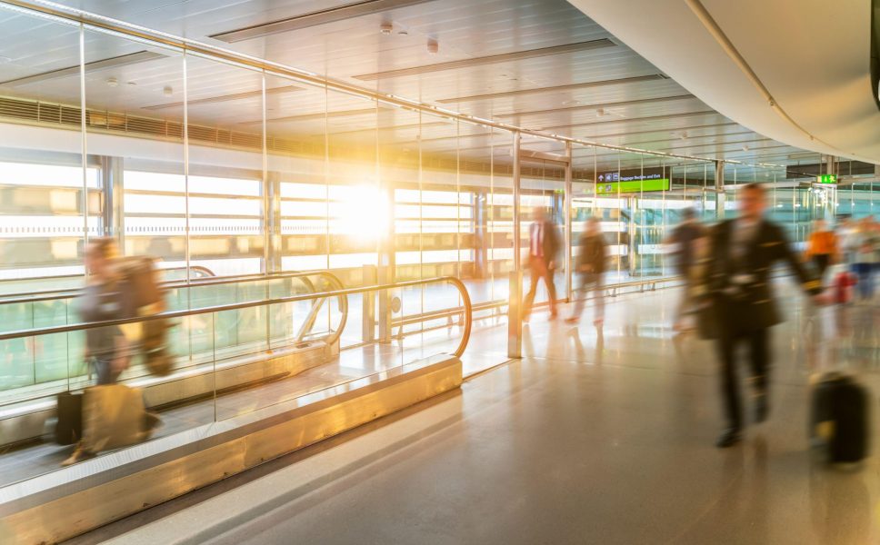 A modern airport terminal corridor with floor-to-ceiling glass windows flooding the space with natural sunlight. The scene captures blurred figures of travellers in motion, creating a sense of bustling activity typical of busy transport hubs. The architecture features sleek metallic railings, polished floors, and contemporary ceiling design with integrated lighting. Through the large windows, aircraft and airport infrastructure are visible on the tarmac outside. Digital flight information displays can be seen in the background, along with directional signage. The motion blur effect emphasises the constant flow of passengers moving through the terminal space, whilst the bright, airy design creates an open and welcoming atmosphere for air travel.