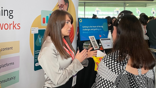 A photograph of the Aurion Learning exhibition stand at the Learnovation 2025 Global Learning Summit. The scene shows two women engaged in conversation at an exhibition stand, with one woman wearing a beige blazer with a pink lanyard and the other in a black and white patterned top.

The setting is within a modern conference venue with distinctive green ceiling features and large windows visible in the background. Behind them is an exhibition booth displaying various learning and development materials, including banners about "Getting to the heart of the big challenges and guiding you through" and references to content and platforms.

The booth features display materials on tablets or digital devices, and there are several other attendees visible in the background, creating the bustling atmosphere typical of professional learning and development conferences. The green stadium seating visible through the windows suggests this may be taking place at a sports venue being used as a conference space.

This image effectively captures the collaborative, professional networking environment that characterises modern learning and development industry events.
